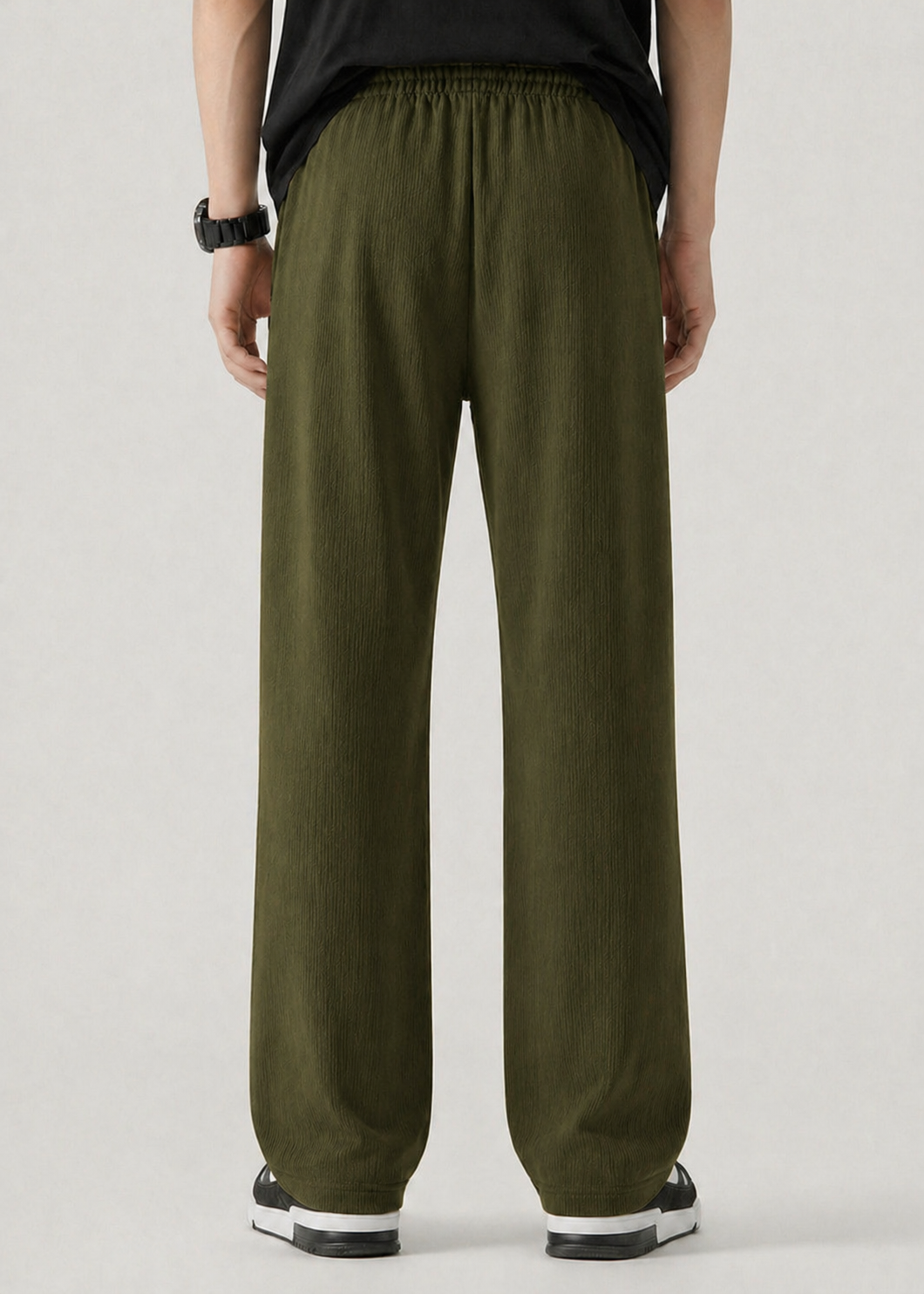 A person shown from the back wearing loose, wide-leg olive green pants with an elastic waistband, paired with a black top and black-and-white sneakers against a light gray studio background.